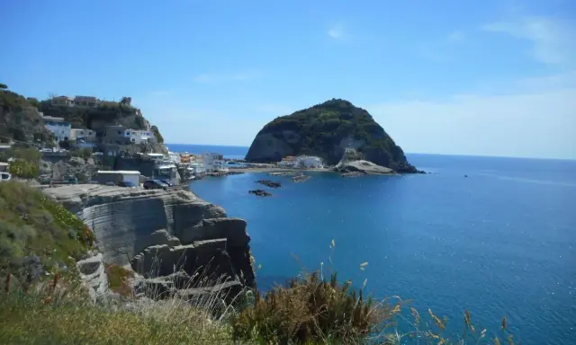 Scogliere a picco sul mare blu, case bianche aggrappate alla costa e un isolotto verde. Un paesaggio che ispira frasi su Ischia.