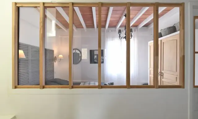 Une verrière en bois sépare deux pièces. On y voit un lit, des volets, une armoire et un lustre.