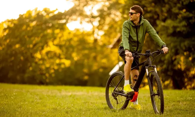 Mężczyzna w zielonej kurtce i okularach przeciwsłonecznych odpoczywa na trawie obok swojego roweru elektrycznego ebike.