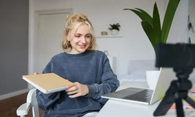 Junge Frau gibt online Nachhilfe, hält ein Notizbuch und lächelt in die Kamera.