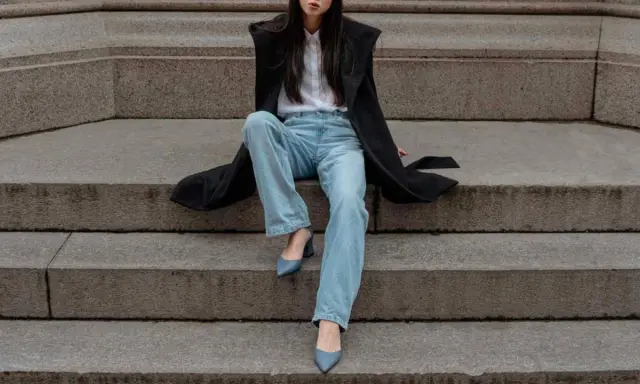 Donna con cappotto nero, camicia bianca e **pantaloni a sigaretta** chiari, indossa **tronchetti** blu.