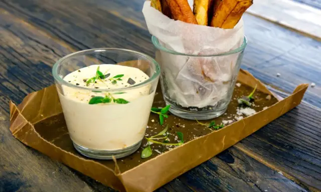 En krämig creme fraiche sås med örter serveras bredvid en glasbehållare med krispiga pommes frites på ett rustikt träbord.