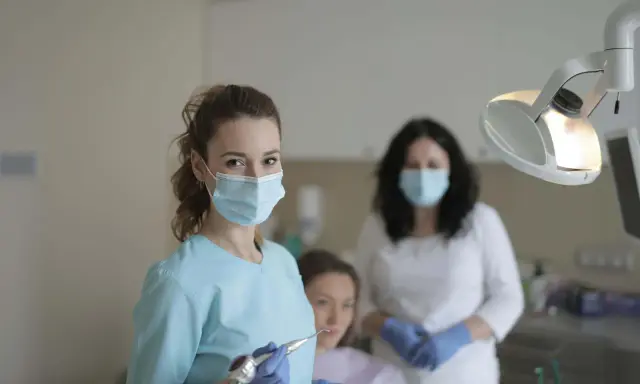 Uśmiechnięta studentka stomatologii w masce i rękawiczkach trzyma wiertło dentystyczne. W tle inne osoby w gabinecie.