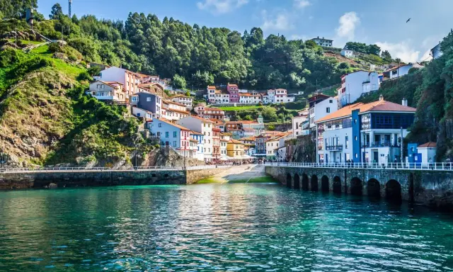 Un pintoresco pueblo costero con **5 villas** coloridas apiladas en una colina verde, frente a un mar turquesa.