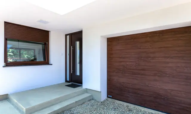 Ingresso con porta scura e avvolgibili marroni, accanto a un'ampia porta garage in legno scuro.