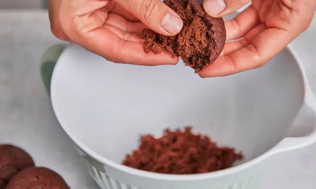 Hände zerbröseln einen Schokoladenkuchen für vegane Desserts. Ein Teil landet in einer weißen Schüssel.