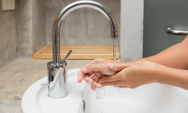 Mains d'une personne se lavant sous un robinet moderne en inox. L'eau coule dans un lavabo blanc.