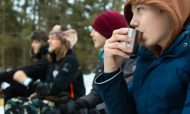 Barn njuter av fika under friluftsdag aktiviteter i snöig skog. En pojke dricker ur en metallmugg.