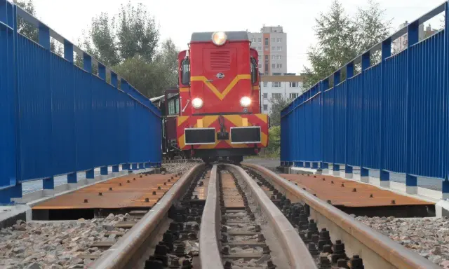 Czerwona lokomotywa na torach wąskotorowych, część górnośląskich kolei wąskotorowych, wjeżdża na most z niebieskimi barierkami.