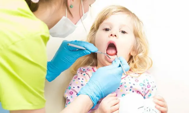 Mała dziewczynka z otwartymi ustami, kiedy z dzieckiem do dentysty. Stomatolog w niebieskich rękawiczkach bada jej ząbki.