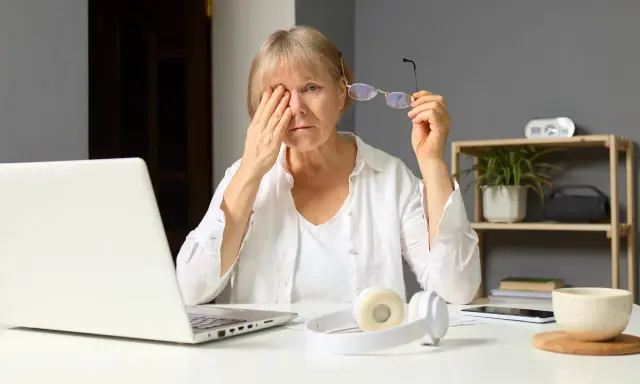 Zmęczona kobieta pociera oko, patrząc na ekran laptopa, który emituje niebieskie światło. Obok leżą okulary i słuchawki.