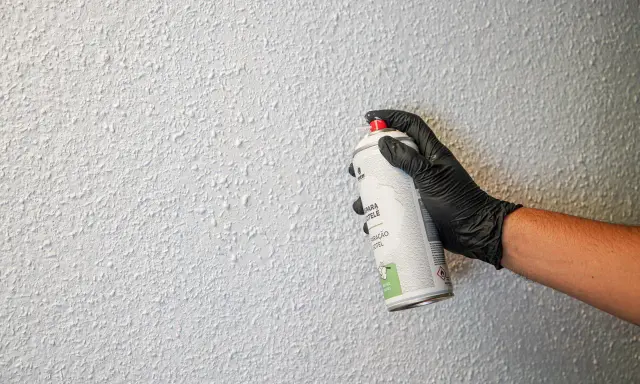 Mano con guante negro aplica spray para reparar gotelé. La pared tiene textura rugosa y blanca.
