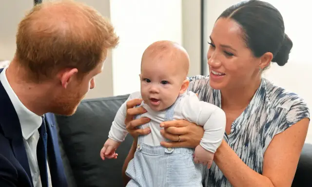 Meghan Markle håller sitt barn, som tittar nyfiket framåt. Prins Harry ser på dem med ett leende.