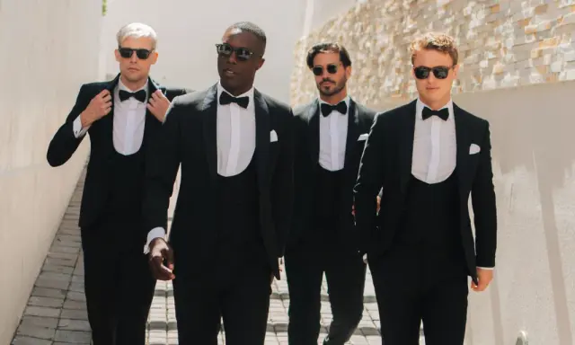 Four men stride confidently in a modern black tie dress code, sporting sharp tuxedos and sunglasses.