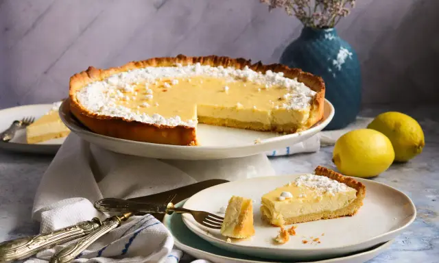 Ein köstliches Zitronentarte Rezept: Eine ganze Tarte und ein Stück auf einem Teller, bestäubt mit Puderzucker. Zwei Zitronen liegen daneben.