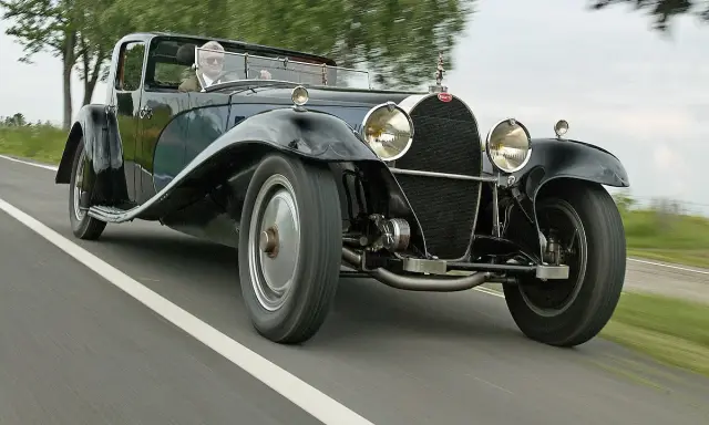 Ein Mann fährt einen glänzenden schwarzen Bugatti Royale auf einer Landstraße. Der Bugatti Royale Preis ist astronomisch, ein Traum für jeden Sammler.