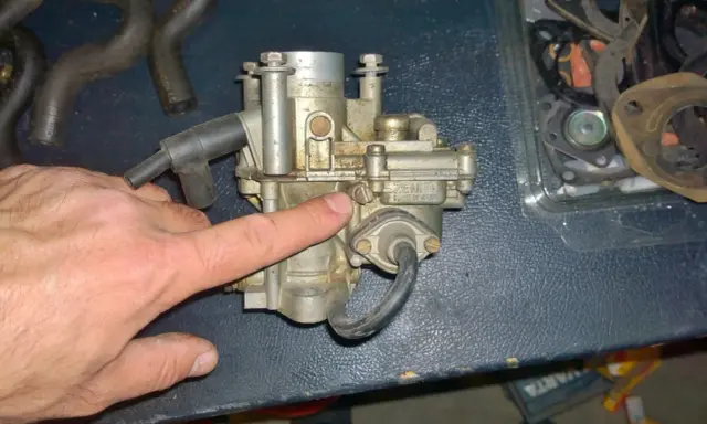 Main d'homme pointant un carburateur Zenith, possible cause d'un moteur 4 temps qui s'étouffe à l'accélération. Pièces de rechange visibles.