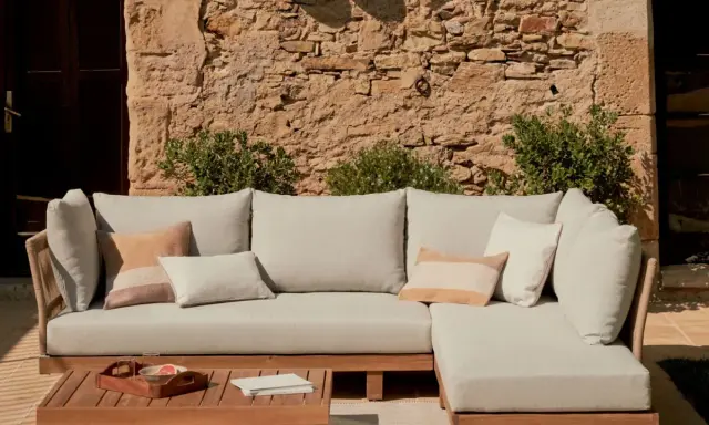 Sofá modular de exterior con cojines en tonos neutros, sobre una mesa de madera. La **decoración estilo natural** se realza con la pared de piedra y la vegetación.