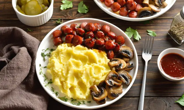 Talerz z puree ziemniaczanym, parówkami i pieczarkami, obok ogórki i sos.