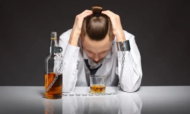 Mężczyzna w kajdanach, przykuty do butelki i szklanki z alkoholem, symbolizuje uzależnienie od piwa i jego niszczycielską siłę.