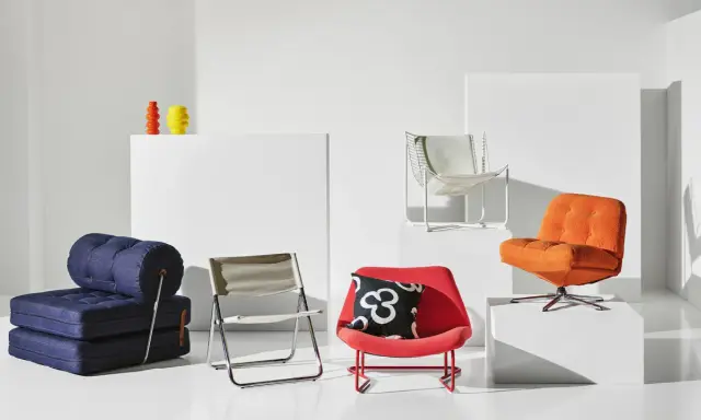 En samling möbler i 70-tals inredning: en blå bäddsoffa, en röd fåtölj med mönstrat kudde, en orange fåtölj och en vit metallstol.