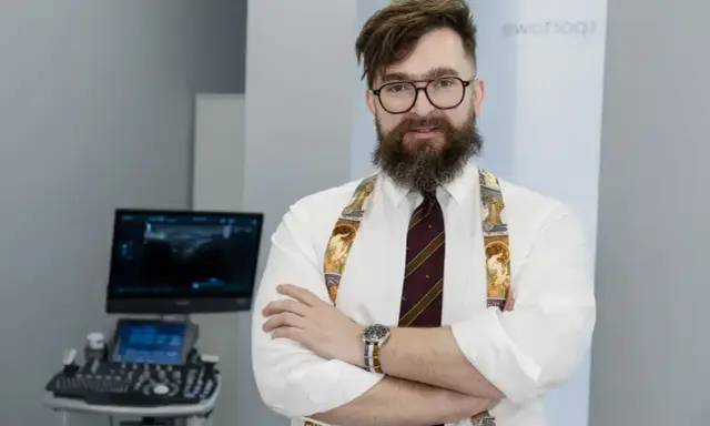 Ortopeda w okularach i ozdobnych szelkach stoi przed aparatem USG.