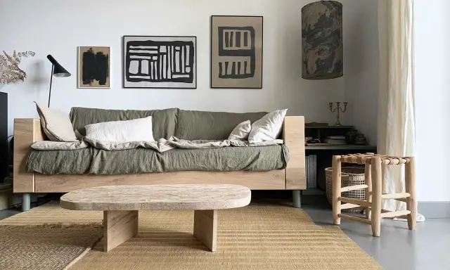 Une idée table basse fait maison en bois brut, posée sur un tapis tressé, devant un canapé vert et des tableaux abstraits.