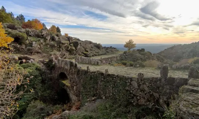 Herbstliche Farben in der Sierra de Gata. Ein alter Steinbogenbrücke, überwuchert von Efeu, führt durch eine felsige Landschaft.