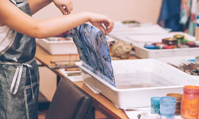 Manos con delantal pintan mármol sobre una tela azul y blanca, sumergiéndola en una bandeja con agua.
