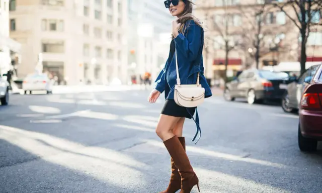 A woman walks down a city street wearing a black dress, a denim jacket, and stylish brown boots.