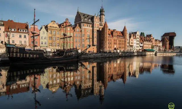 Gdańsk atrakcje: zabytkowy żaglowiec cumuje przy Motławie, w tle kolorowe kamienice i Żuraw. Odbicia budynków w wodzie.