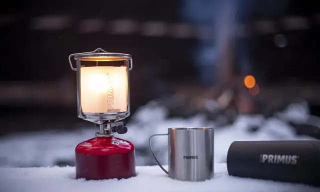 En varm primus lampa lyser upp snön bredvid en mugg och en termos. I bakgrunden syns en lägereld.