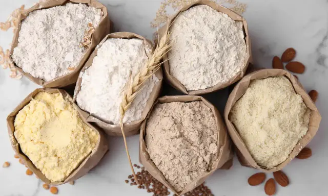Six sacs en papier remplis de différentes farines, dont une farine de blé CRC. Amandes et grains parsèment le fond marbré.