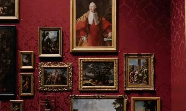 A gallery wall displays classical paintings in ornate frames against a rich red damask background. This collection showcases the best way to invest in art, with portraits and landscapes hinting at historical narratives.