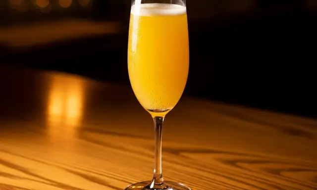 Ett glas med en gyllene mimosa drink, bubblig och uppfriskande, står på ett träbord i varmt ljus.