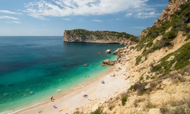 Türkisblaues Wasser trifft auf Kiesstrand in einer malerischen spanischen Atlantikbucht. Felsen und grüne Vegetation säumen die Küste.