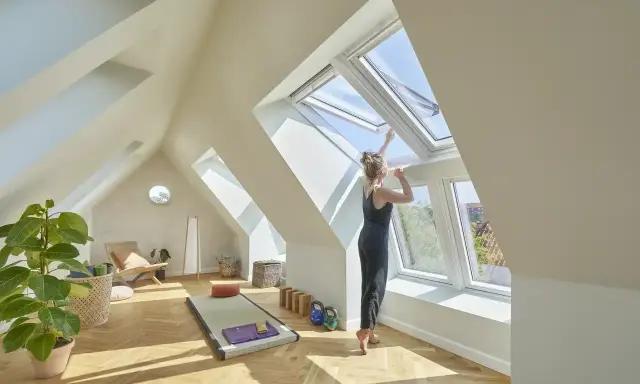 Frau öffnet Dachfenster in hellem Dachboden-Yoga-Raum. Hier darf man Dachboden als Wohnraum nutzen und schafft sich eine Oase.