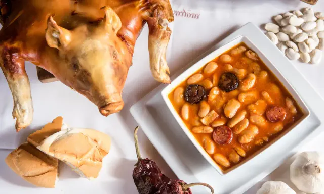 Cochinillo asado segoviano, crujiente y dorado, acompañado de un plato de judiones con chorizo y morcilla, pan y ajos.
