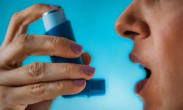 Person nutzt Asthmaspray vor oder nach dem Sport. Die Hand hält das blaue Gerät, der Mund ist geöffnet.