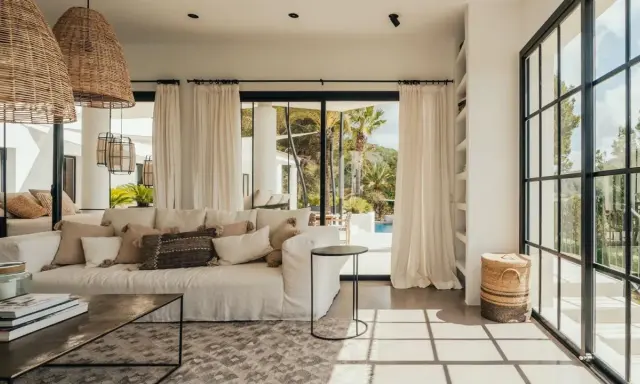 Arredare una casa al mare: divano bianco, cuscini naturali, lampade in rattan e vista su piscina e palme.