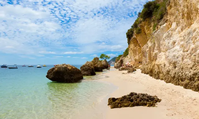 Sonniger Setúbal Strand mit türkisfarbenem Wasser, Felsen und Booten im Hintergrund. Eine kleine Gruppe entspannt sich unter einem Sonnenschirm.