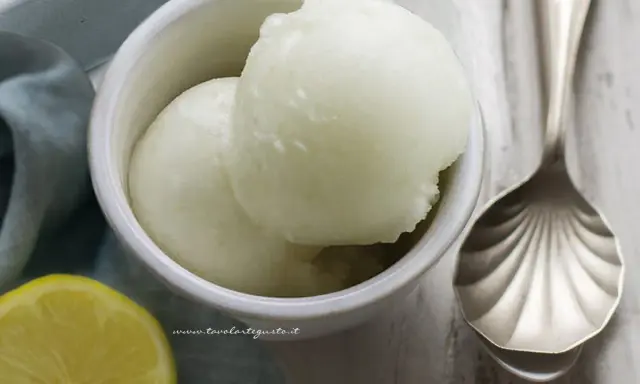 Coppetta con due palline di sorbetto al limone senza albume, accanto una fetta di limone e un cucchiaio decorato.