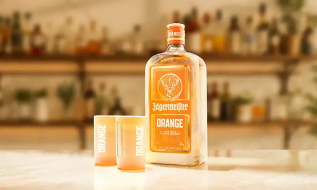 Butelka Jägermeister Orange i dwa kieliszki z napisem "ORANGE" na blacie. Idealny zestaw na letnie wieczory.