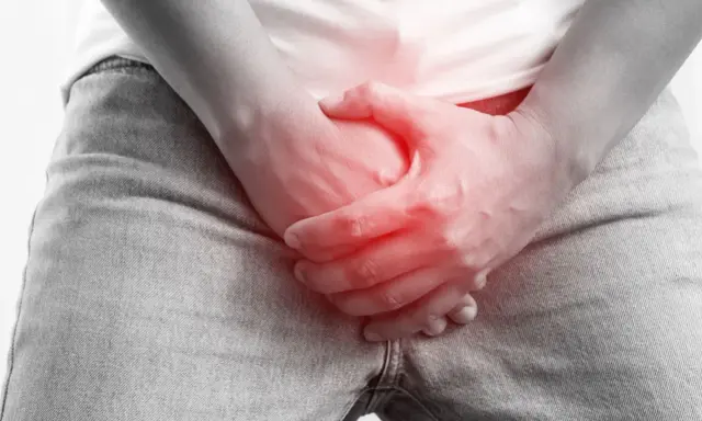Dłoń obejmuje krocze z czerwonym podświetleniem, sugerując ból lub uraz, jak złamanie prącia.