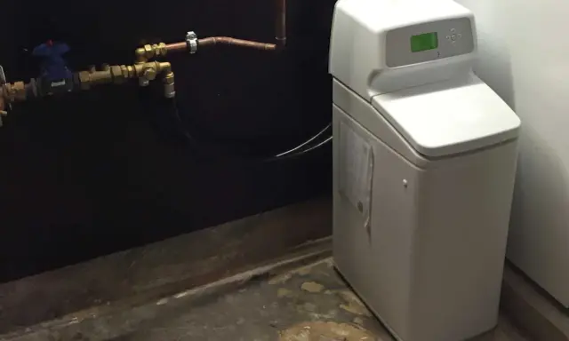 Appareil blanc avec écran vert, connecté à des tuyaux en cuivre. C'est un adoucisseur d'eau potable pour une maison.
