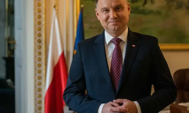 Prezydent Andrzej Duda w garniturze, z polską flagą w tle, czeka na moment, kiedy prezydent podpisze emerytury stażowe.