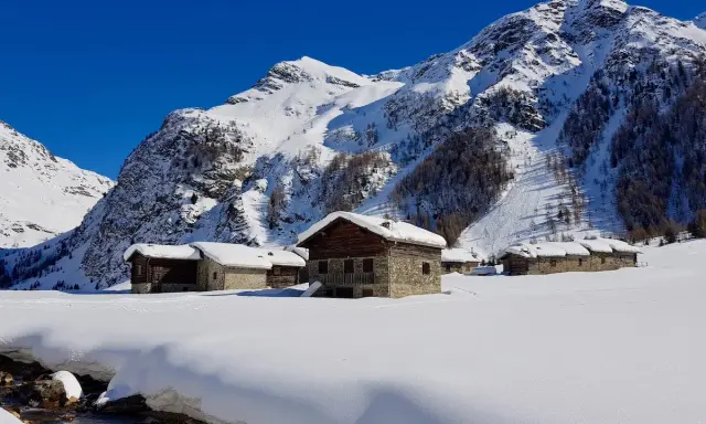 Case innevate nel Val di Rezzalo, con baite in pietra e legno. Per sapere val di rezzalo come arrivare, segui il ruscello che scorre tra la neve.
