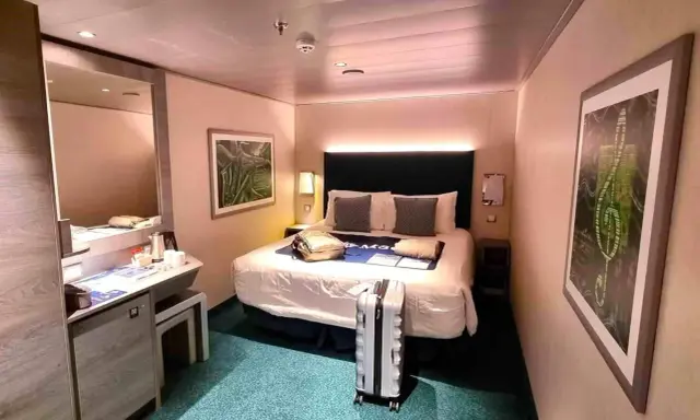 Interno di una royal caribbean cabine con letto matrimoniale, specchio, scrivania e valigia pronta per il viaggio.