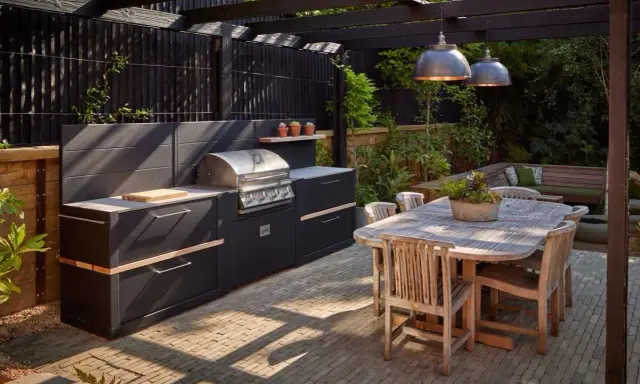 Nowoczesny grill w ogrodzie, z czarną zabudową i drewnianym stołem, zaprasza do wspólnego biesiadowania.