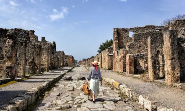 Una donna con cappello di paglia cammina lungo una strada lastricata di Pompei, tra antiche rovine. Un'esperienza unica per scoprire cosa vedere a Pompei.